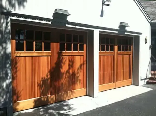 Two wooden garage doors with upper windows on a white house in sunlight
