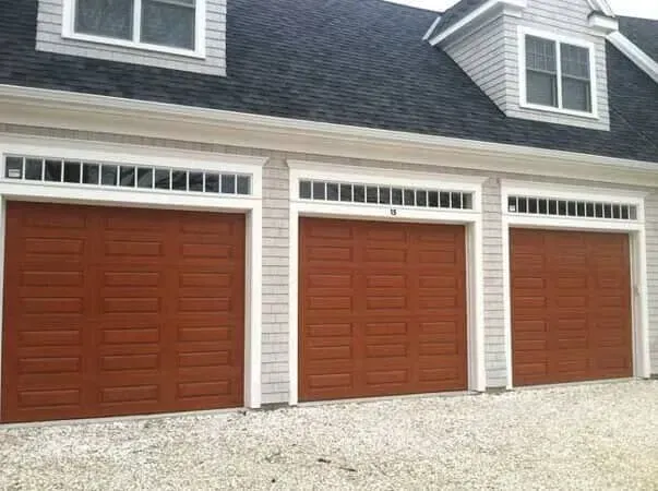Three brown garage doors on a white house with gray shingles and gravel driveway