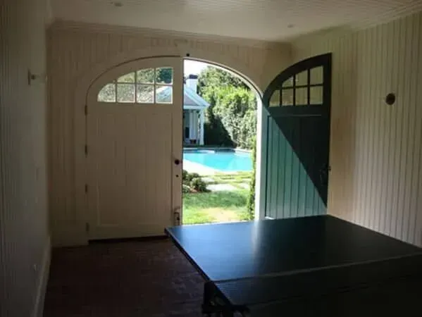 Open double doors to a sunny backyard with a pool, viewed from a dim room with a black table.
