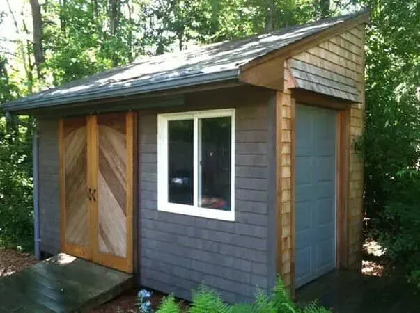 Small gray shed with wooden doors and white window in a wooded backyard.