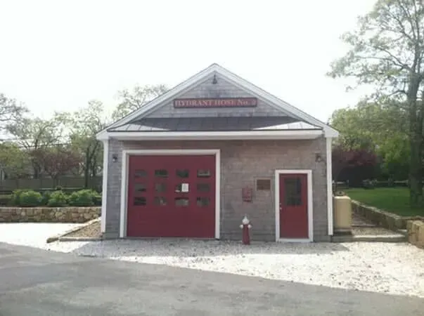 Small red-brick fire station with a red garage door and side entrance, surrounded by trees.