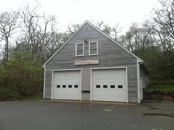 Gray two-bay garage with white doors in a wooded lot on an overcast day