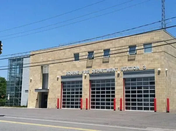 Exterior of a beige fire station with red garage doors and a glass tower under a clear blue sky