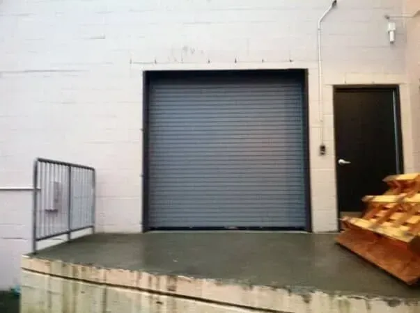 Closed gray roll-up door on a white warehouse wall beside a dark doorway and stacked pallets on a loading dock.