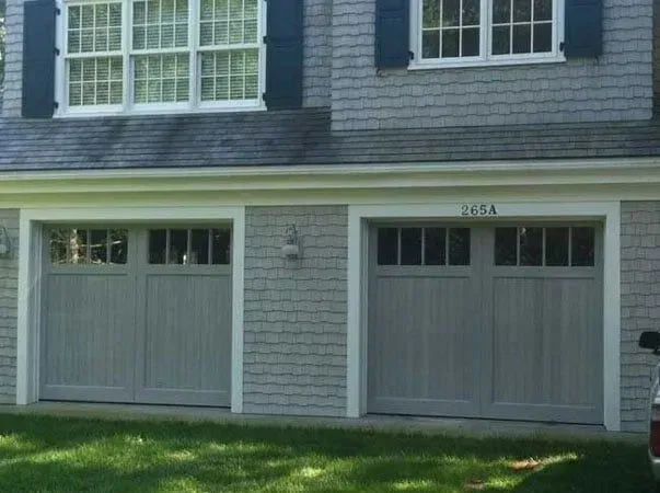 Two gray garage doors beneath a gray house facade with white-trimmed windows and green lawn
