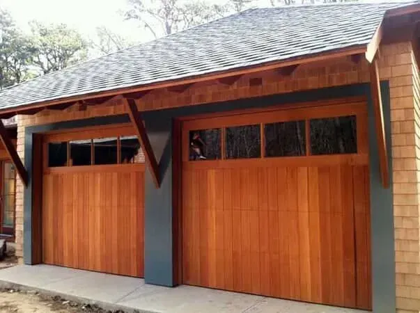 Wooden double garage doors with dark trim beneath a shingled roof on a brick house