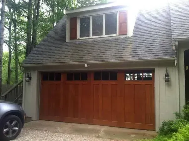 Brown wooden garage door on a house with gray siding and dormer windows, surrounded by trees