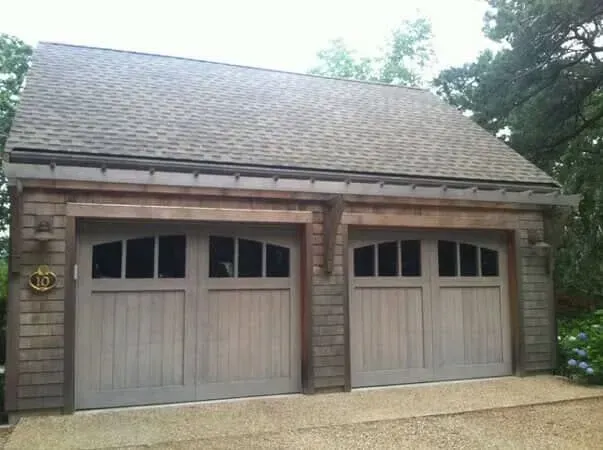 Double garage with two tan wooden doors and a shingled roof, viewed from the driveway.