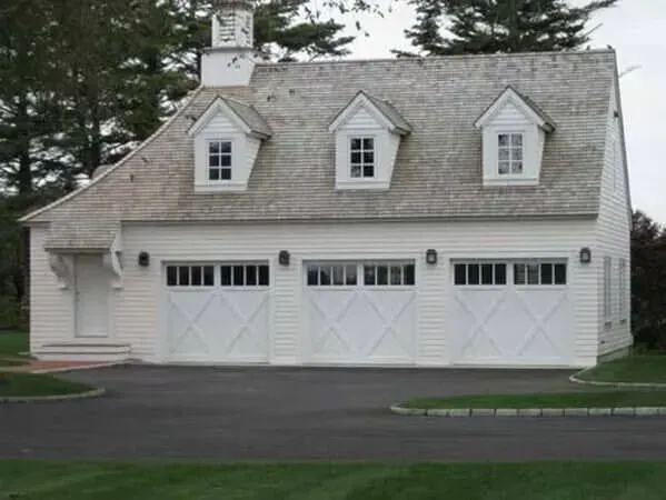 White three-car garage with dormer windows and black roof, set beside a driveway and trees.