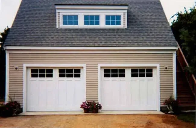 Two white garage doors on a beige house with a gray shingled roof and small dormer windows.