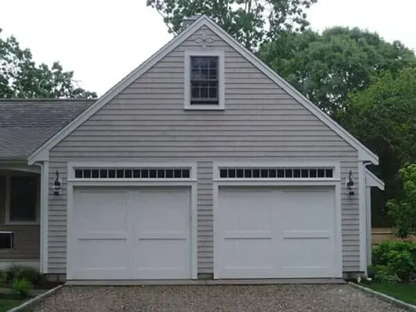 Two-car gray garage with white doors and a peaked roof surrounded by trees