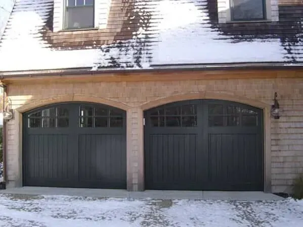 Two dark green garage doors on a brick house with snow on the roof and driveway