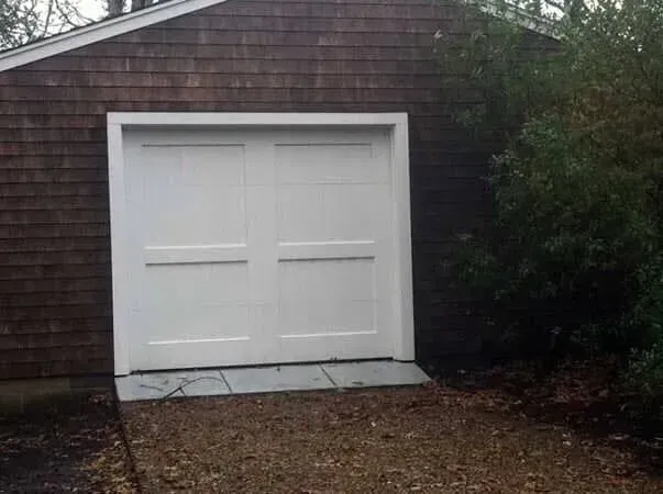 White double garage door on a brown shingle house, with a concrete ramp and gravel driveway.