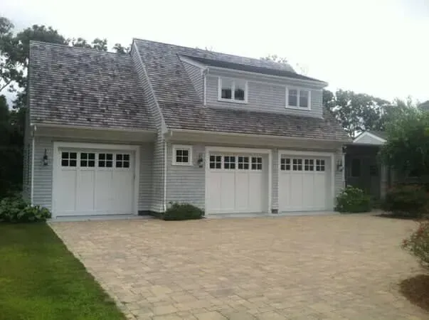 Gray house with three garage doors and a wide tan brick driveway under an overcast sky