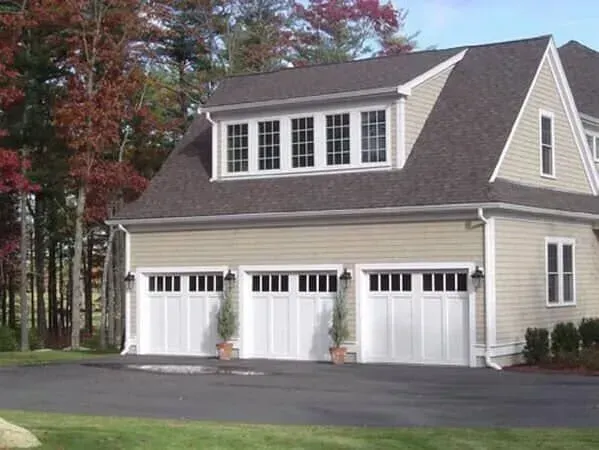 Beige two-story house with three white garage doors and autumn trees in the background