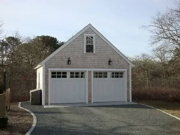 Two-car garage with white doors and gray shingle siding at the end of a gravel driveway