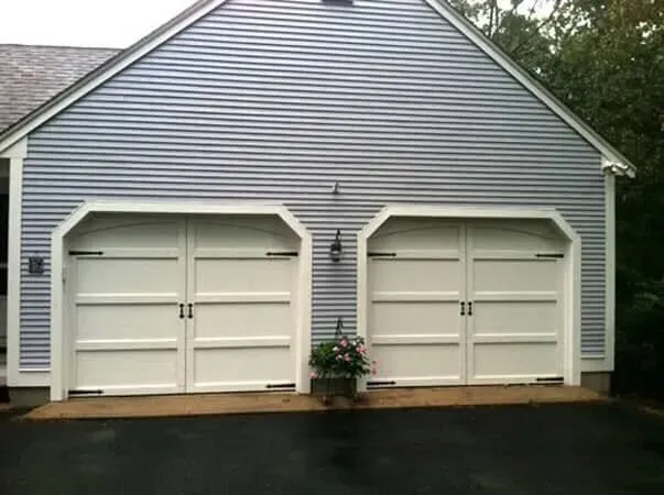Two white garage doors on a gray house with a small flower pot in front and a wet driveway.