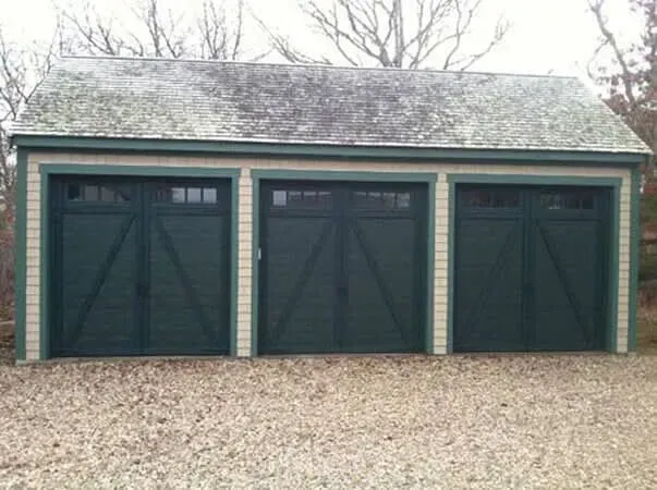 Three dark green garage doors on a brick building with a gravel driveway and bare trees behind it