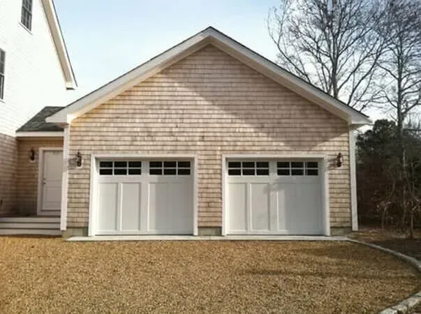 Two-car garage with white doors and beige siding, attached to a house with a gravel driveway.