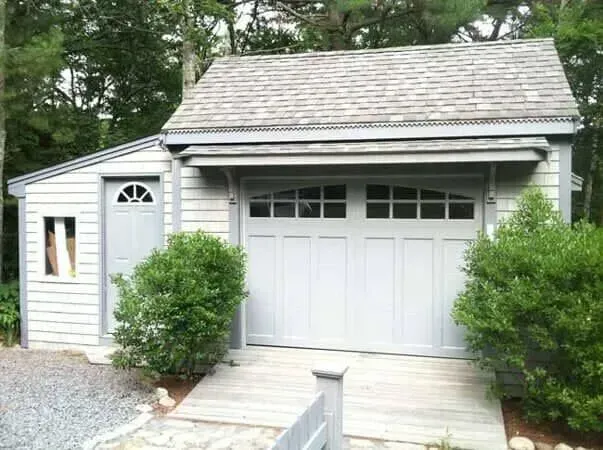 Small white garage with double doors and a side entry door, surrounded by shrubs and trees.
