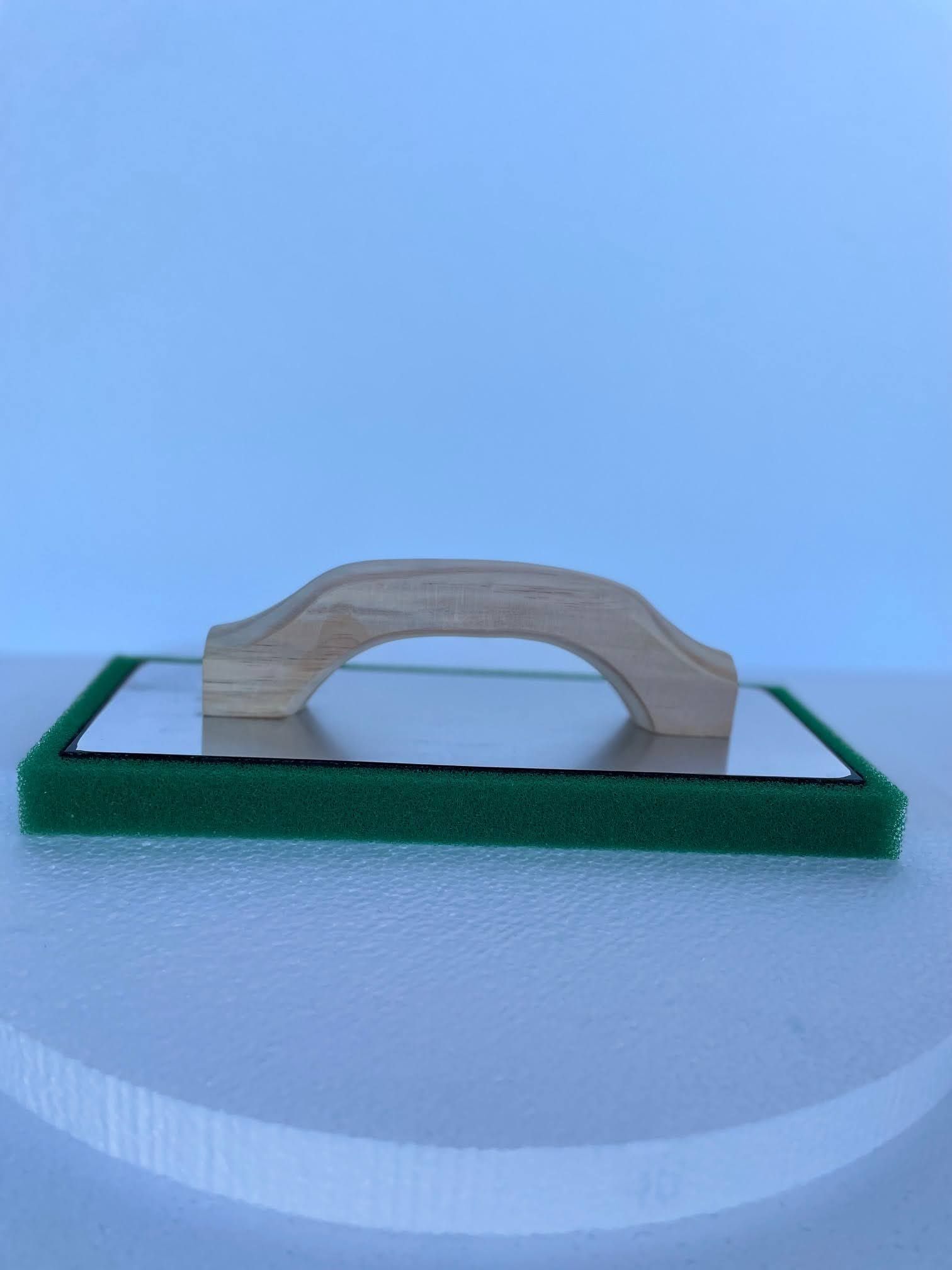A green sponge with a wooden handle is sitting on a white surface.