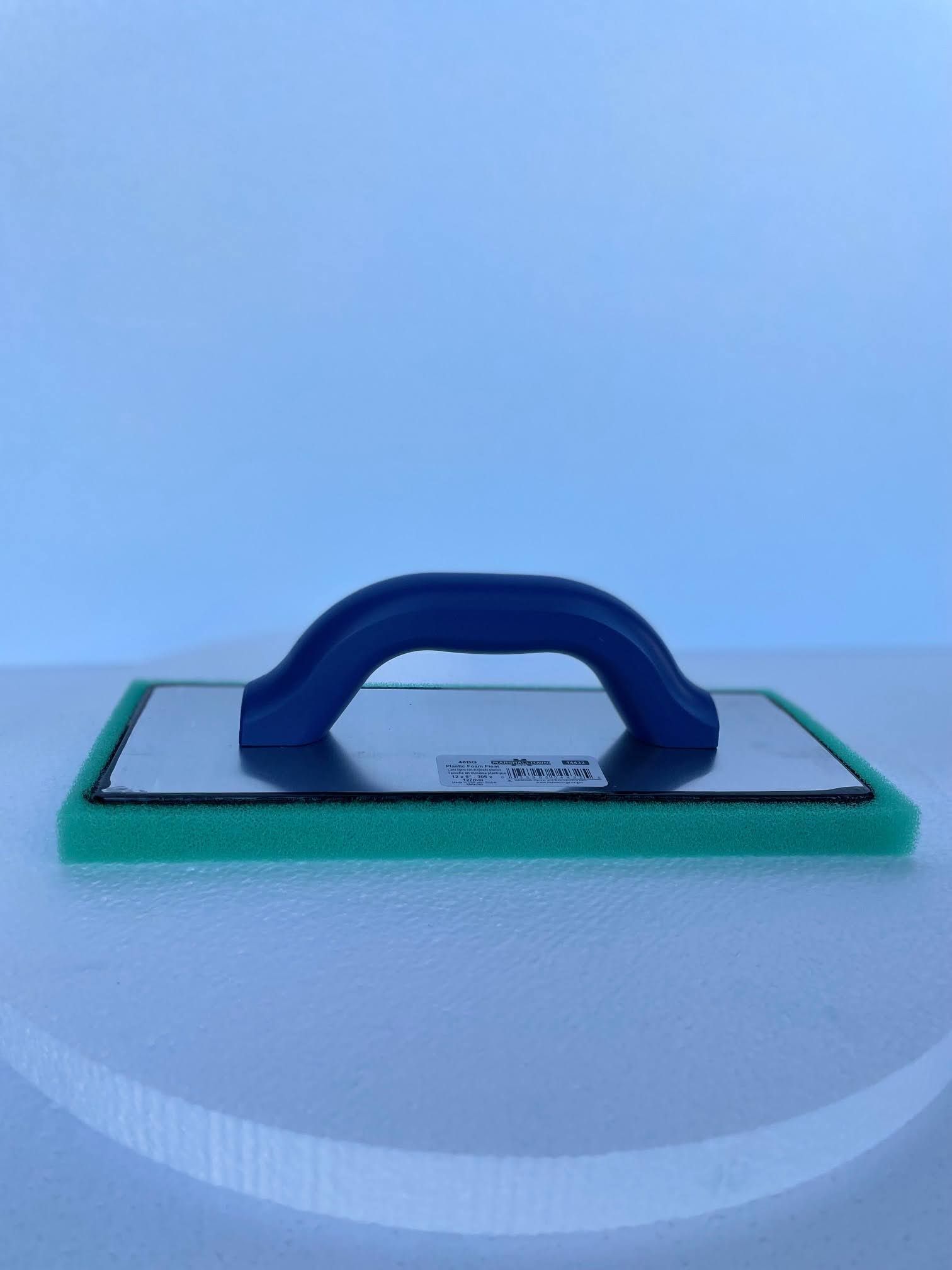 A green sponge with a blue handle is sitting on a white surface.