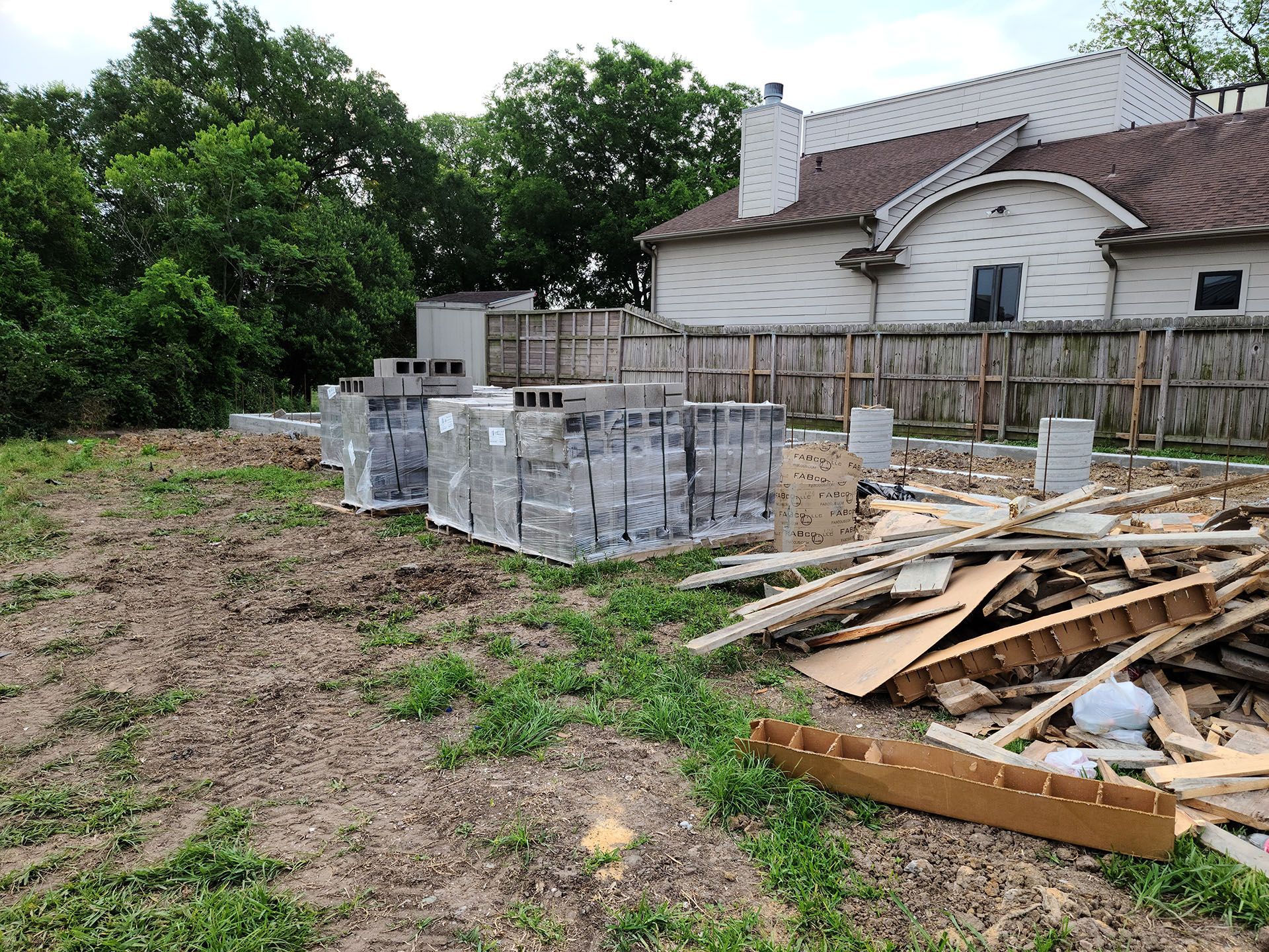 A pile of bricks and wood is sitting in a field in front of a house under construction.