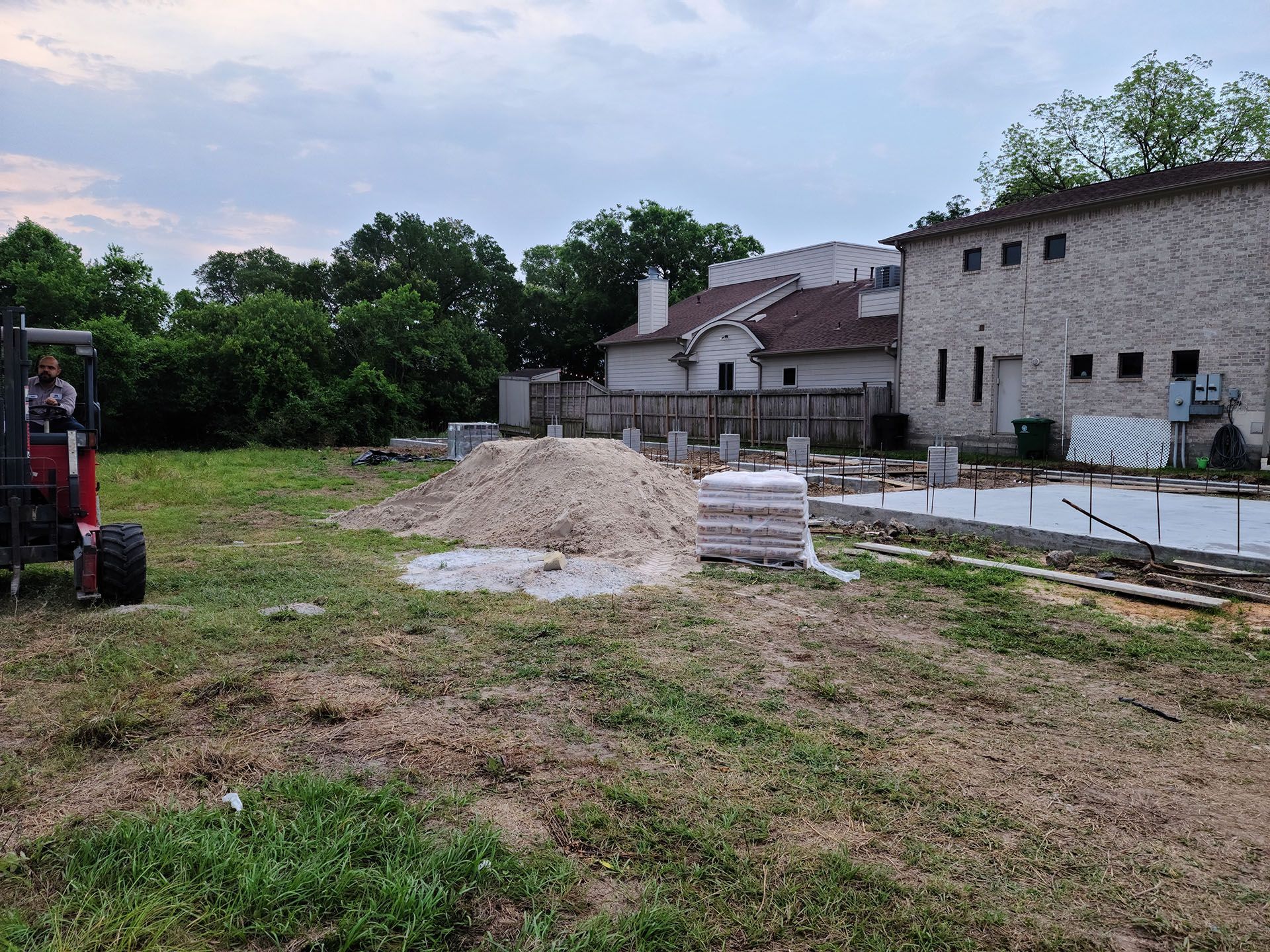 A man is driving a forklift in a grassy field in front of a building under construction.
