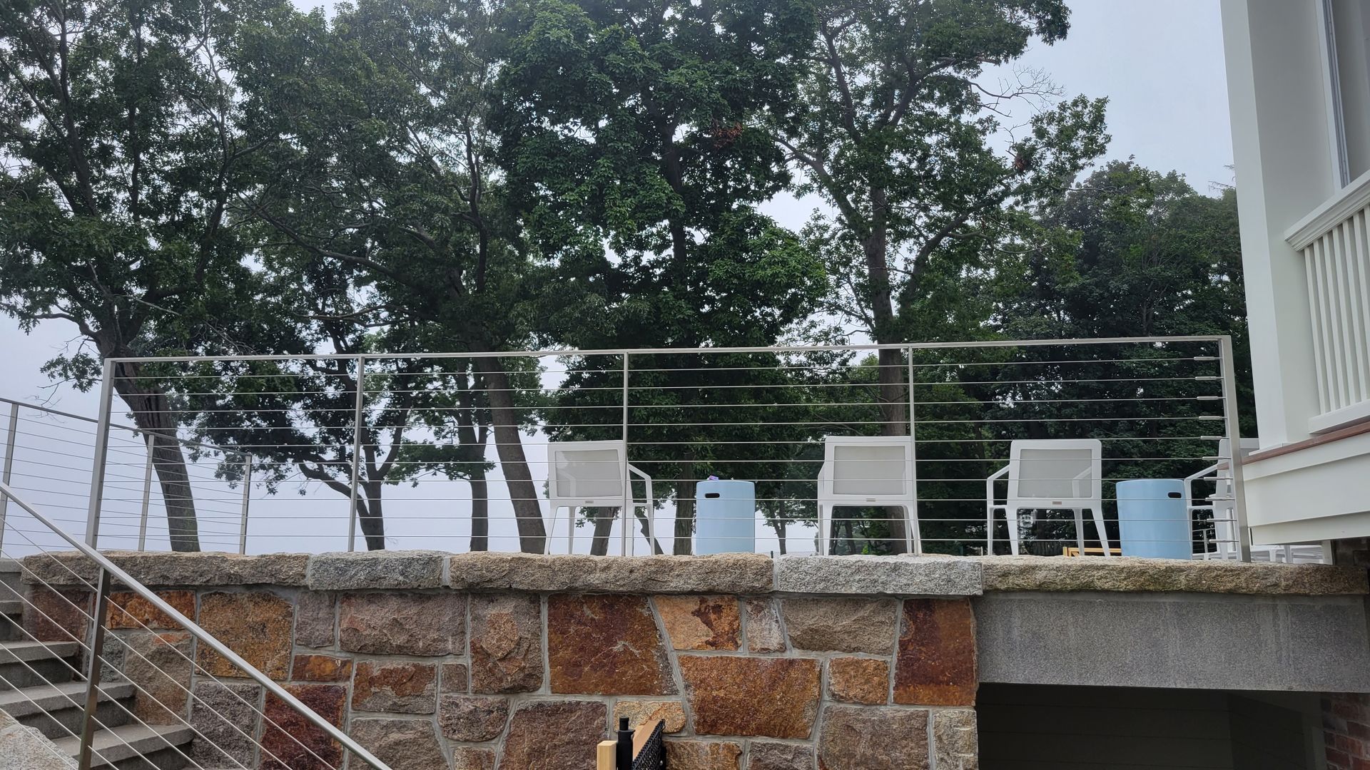 A stone wall with stairs leading up to a deck with chairs and trees in the background.