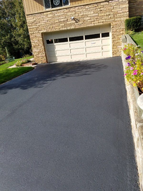 Freshly paved dark driveway leading to a beige garage door beside a stone house and flowers