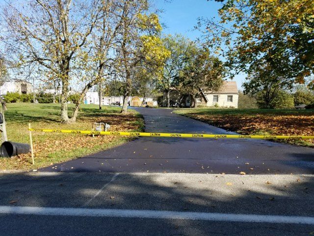 Yellow caution tape blocks a paved park path under leafless trees, with buildings in the background.