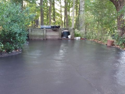 Freshly poured dark concrete patio in a wooded backyard with a hot tub and garden tools at the far end