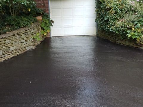 Wet black driveway leading to a white garage door, bordered by stone walls and greenery