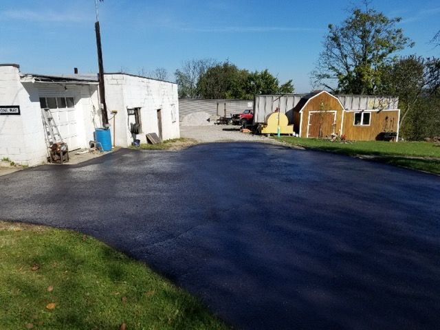 Paved driveway beside white buildings, sheds, and a grassy yard on a sunny day