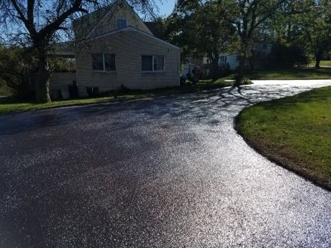 Wet driveway curving past a beige house with trees and sunlight reflecting off the pavement