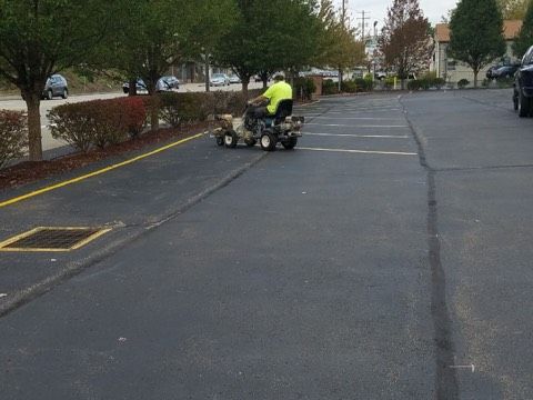 Person on a lawn tractor mowing a parking lot edge beside trees and parked cars