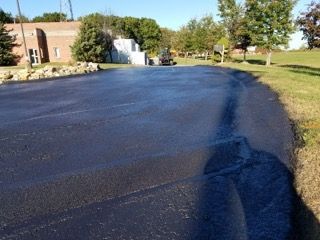 Freshly paved dark asphalt driveway beside a brick building and grassy lawn on a sunny day