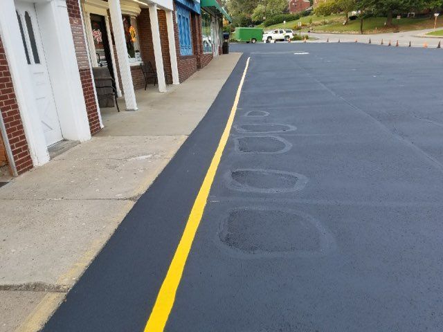Storefront sidewalk beside a fresh yellow curb line and newly paved black parking lot.