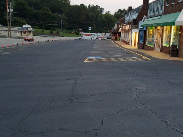 Empty parking lot beside lit storefronts at dusk, with trees and a road in the background.