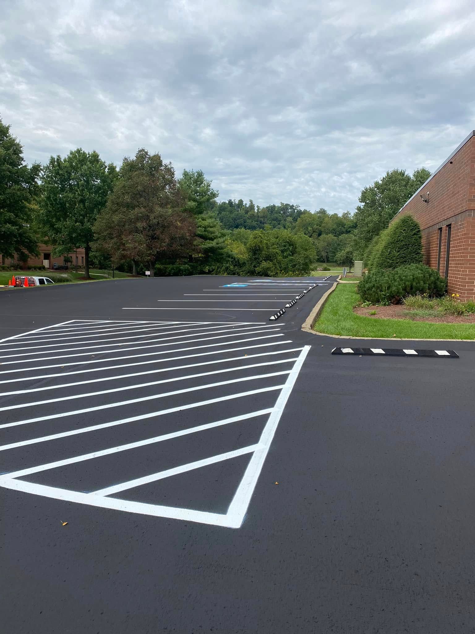 Empty asphalt parking lot with white diagonal markings beside a brick building and trees under cloudy sky