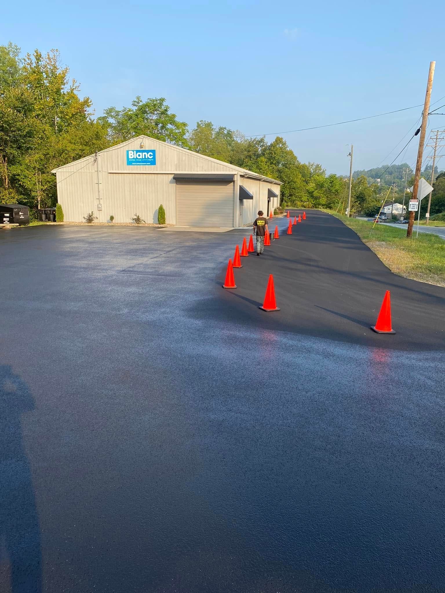 Orange cones line a newly paved driveway beside a tan industrial building in daylight.