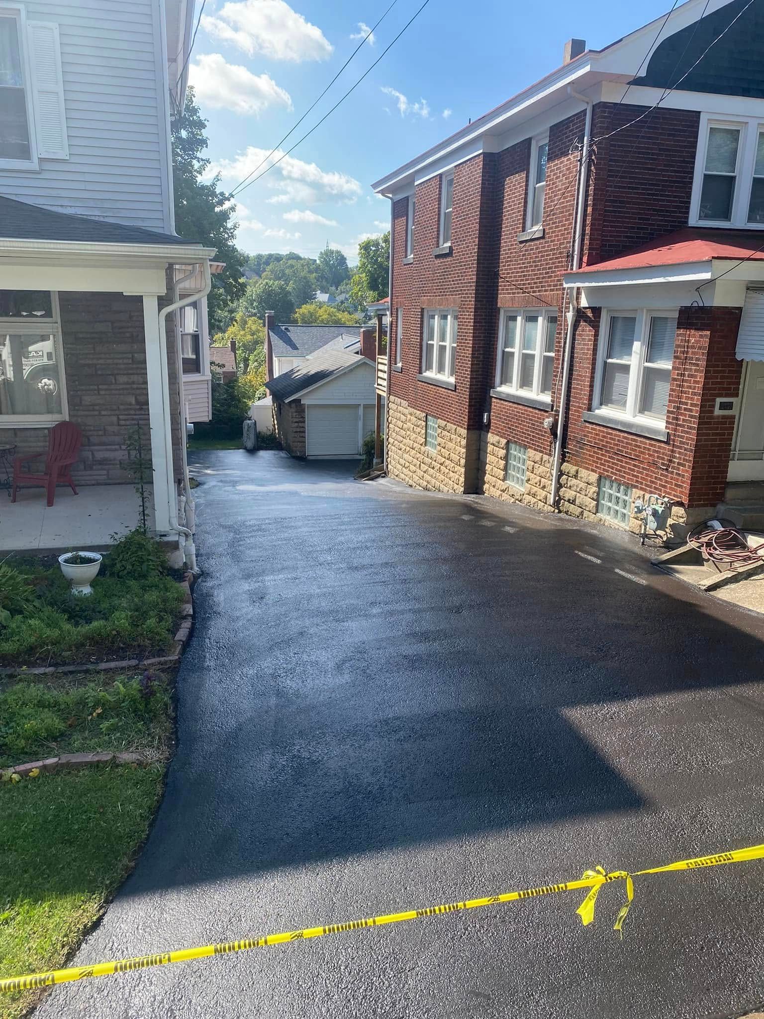 Fresh black asphalt driveway between houses on a sloped residential street, marked with yellow tape in front.