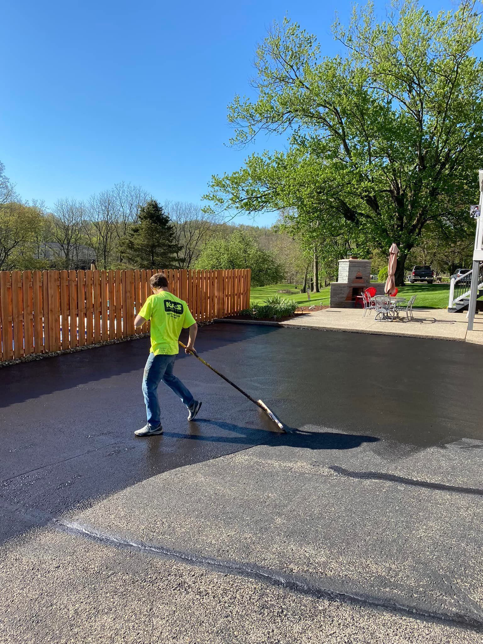 Worker raking gravel on a driveway beside a wooden fence and trees on a sunny day