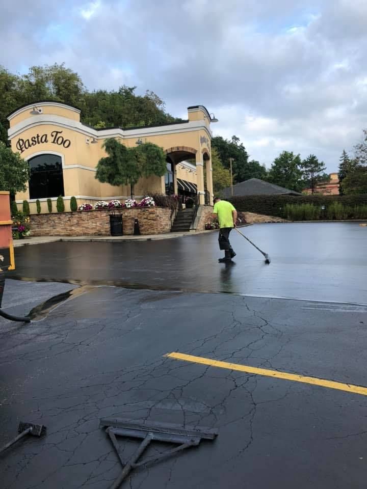 Worker sealing wet parking lot beside a Spanish-style building with a yellow line and hose on the ground