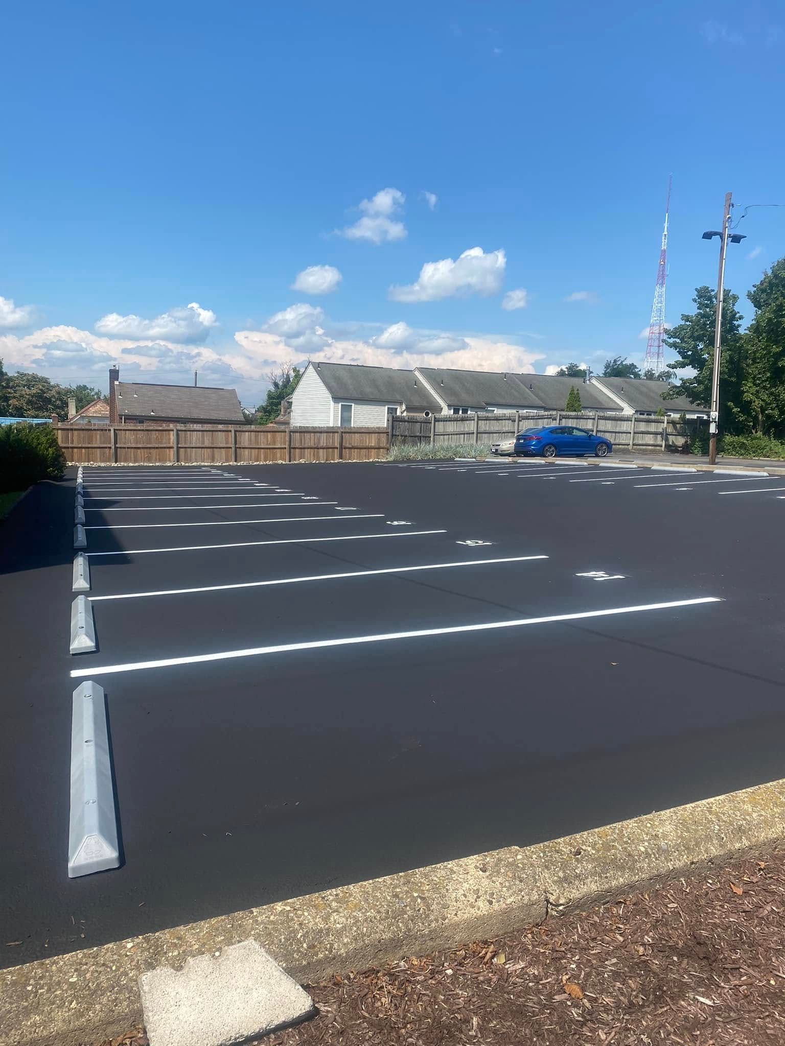 Empty paved parking lot with white lines, a few buildings in the distance, and a blue sky with clouds.