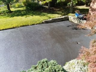 Backyard patio with a dark paved surface, green lawn, shrubs, and a low stone border