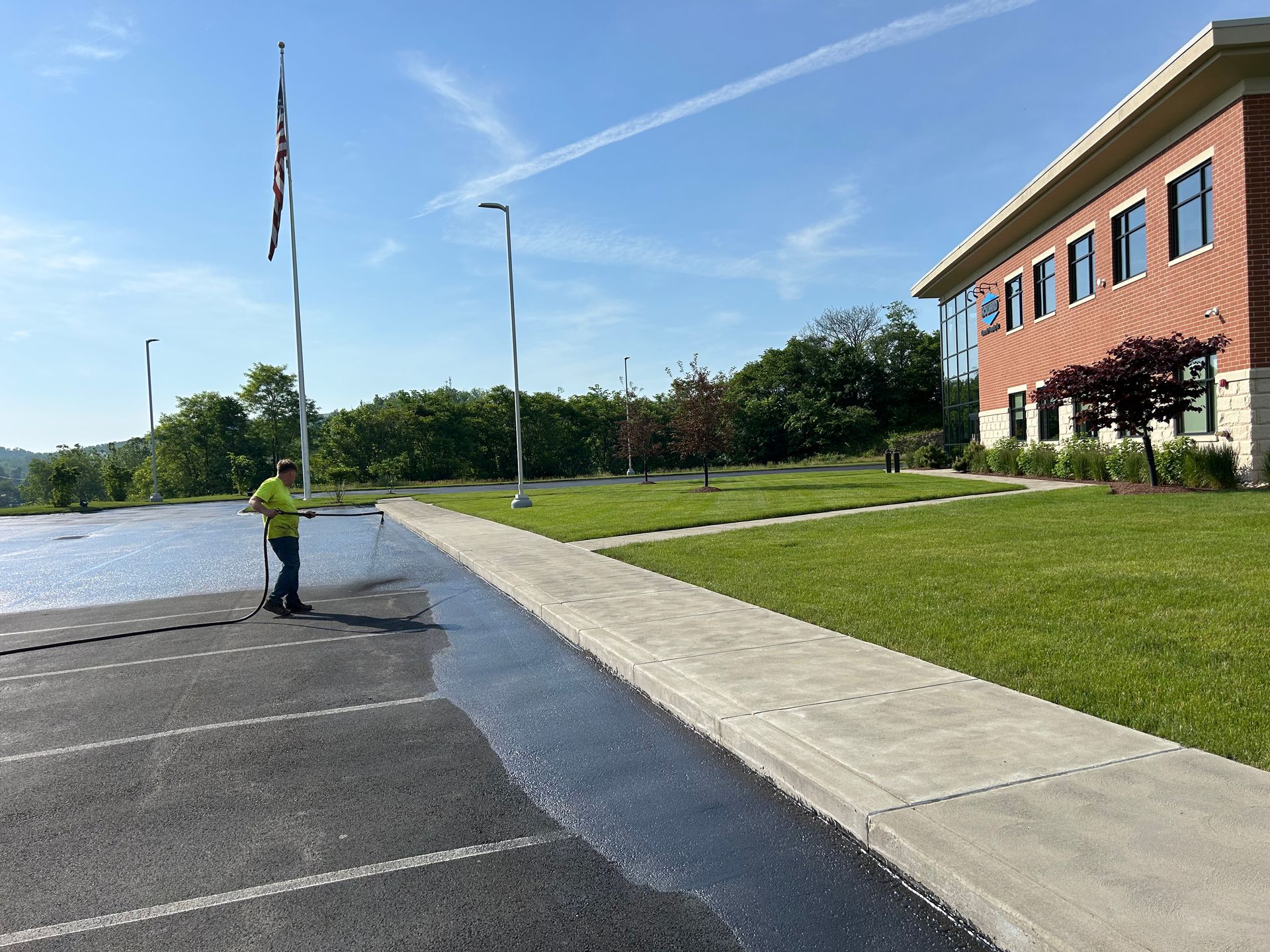 Parking lot and sidewalk beside a red brick building, with a flagpole, grassy lawn, and a person near the water.