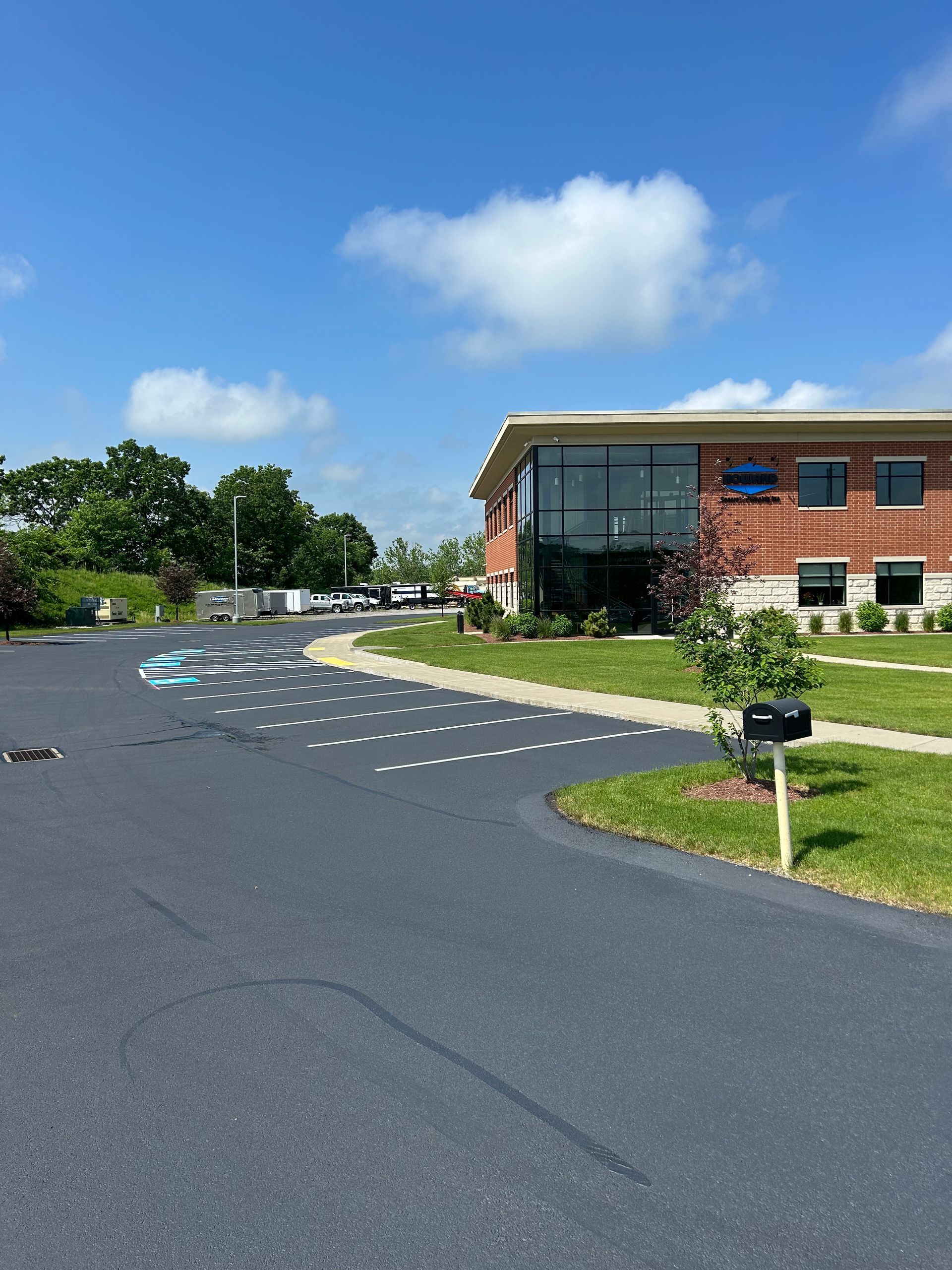 Brick office building beside a curved asphalt road with painted markings, grass, trees, and a mailbox under blue sky
