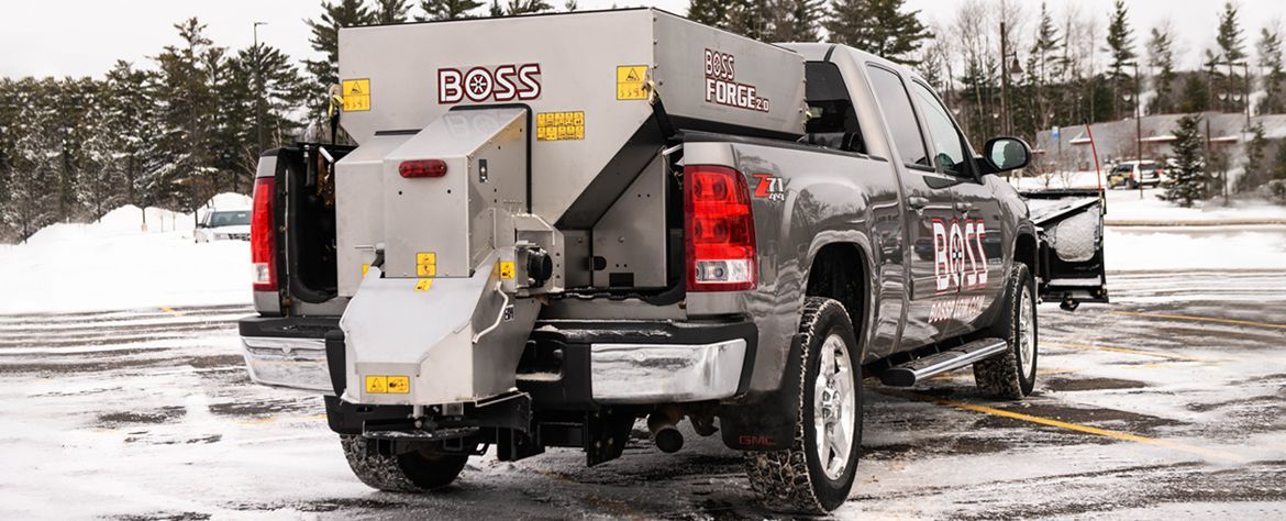 Gray pickup truck with salt spreader in snowy parking lot.