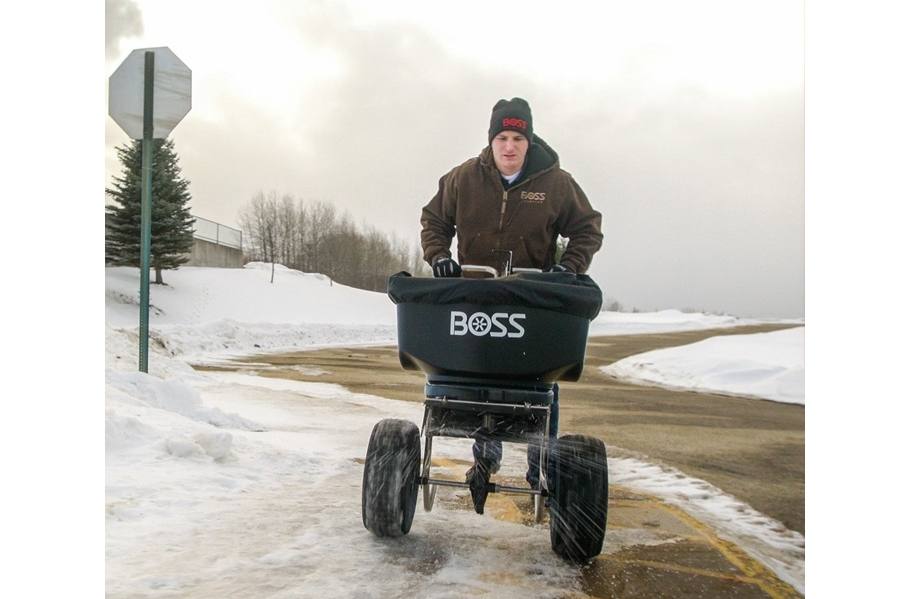 Person spreading salt with a BOSS spreader on a snowy sidewalk near a road and sign.